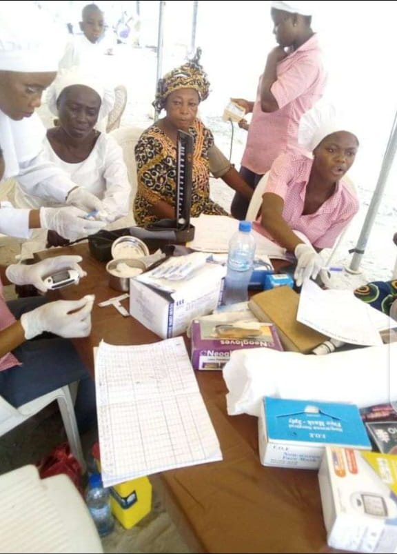 Nurses in gloves taking readings with a blood pressure monitor and testing kit while a woman waits to be seen.